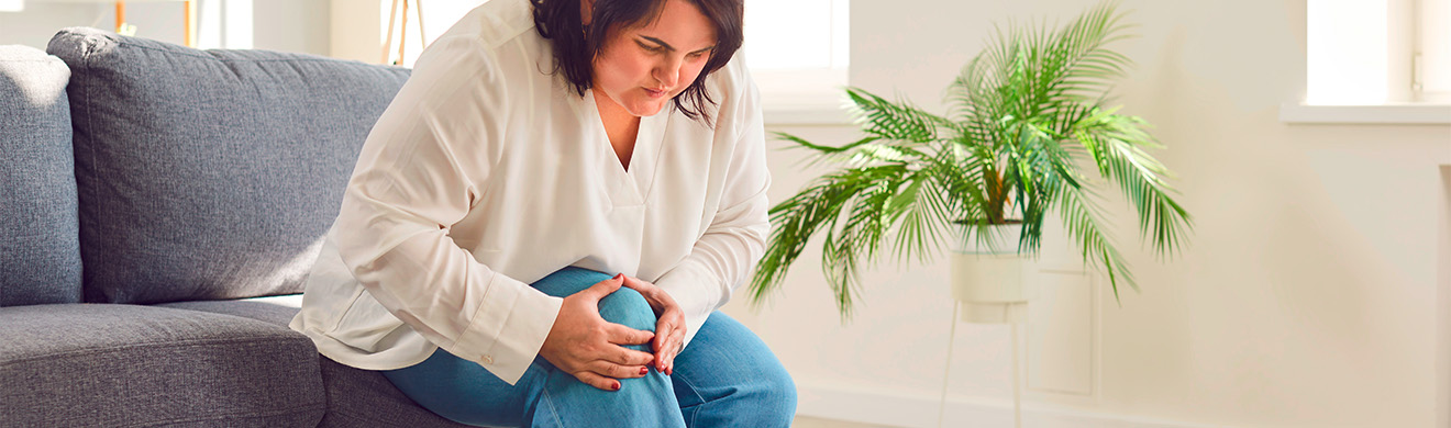 Junge übergewichtige Frau sitzt auf Sofa und hält schmerzendes Knie, Ausdruck von Beschwerden bei chronischen Gelenkschmerzen im Alltag